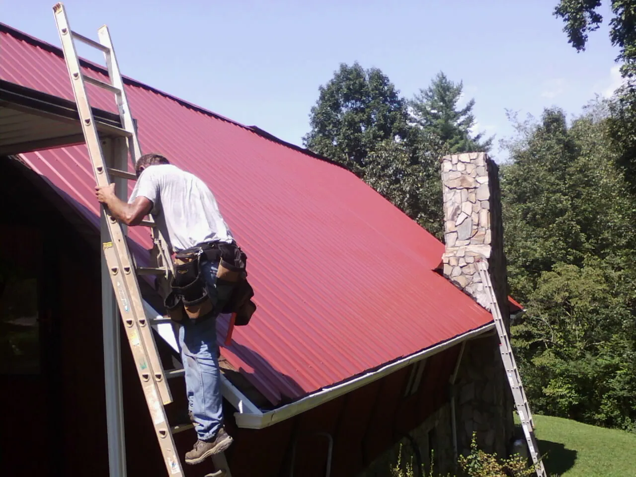 Roofer on a ladder beside a red metal roof and stone chimney — Soffit & Fascia Repair by Roofing Brothers in Landisburg